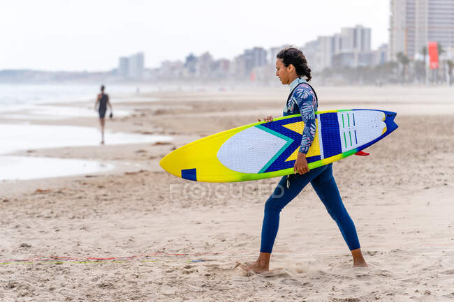 Atleta feminina étnica alegre em wetsuit com kiteboard andando na costa arenosa contra pipa inflável — Fotografia de Stock