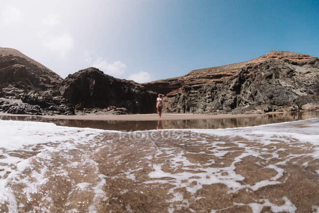 Anonymous female tourist in swimsuit touching forehead between mount and foamy ocean under cloudy blue sky — Stock Photo