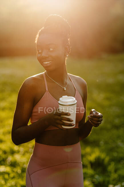 Délicieuse athlète afro-américaine en vêtements de sport debout avec tasse de boisson dans le parc le soir et regardant ailleurs — Photo de stock