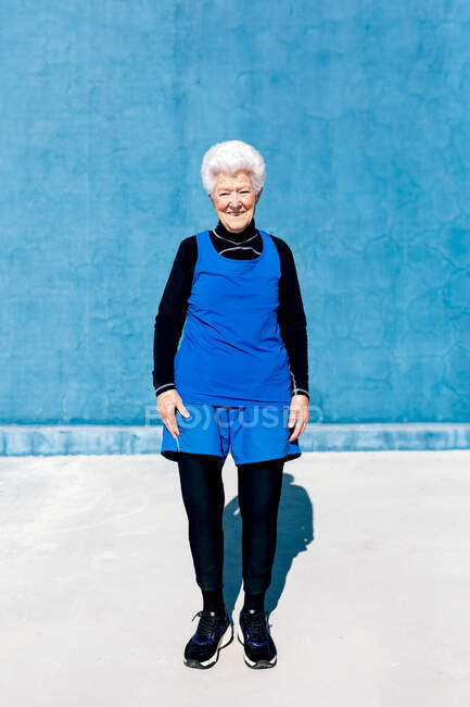 Все тело счастливой зрелой женщины в спортивной одежде, стоящей напротив синей стены в открытом спортивном центре и смотрящей в камеру с улыбкой — стоковое фото