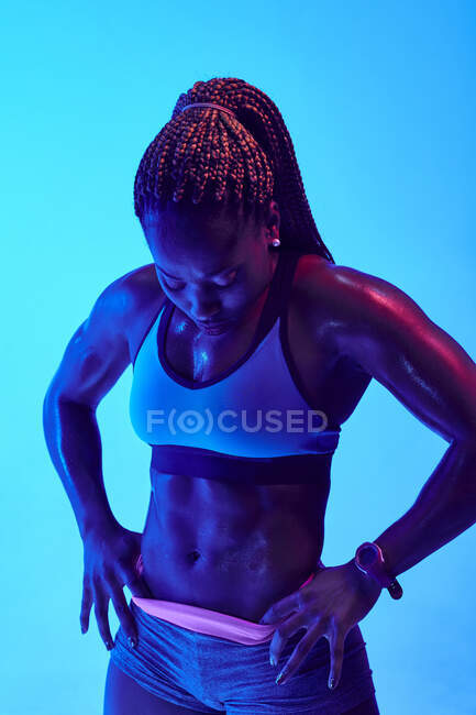 Deportista femenina afroamericana decidida en ropa deportiva tocando los muslos y mirando hacia adelante durante el descanso del entrenamiento con luz de neón - foto de stock