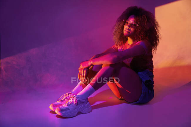 Full body young attractive African American female in shorts and trendy sunglasses sitting on floor in neon lights and embracing knees while looking at camera — Stock Photo