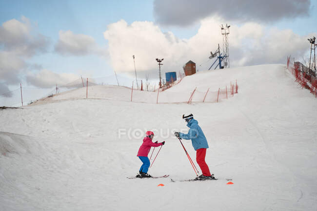 Corpo inteiro pai sem rosto em sportswear quente e capacete ensinando criança a esquiar ao lado de encosta neve colina no inverno estância de esqui — Fotografia de Stock