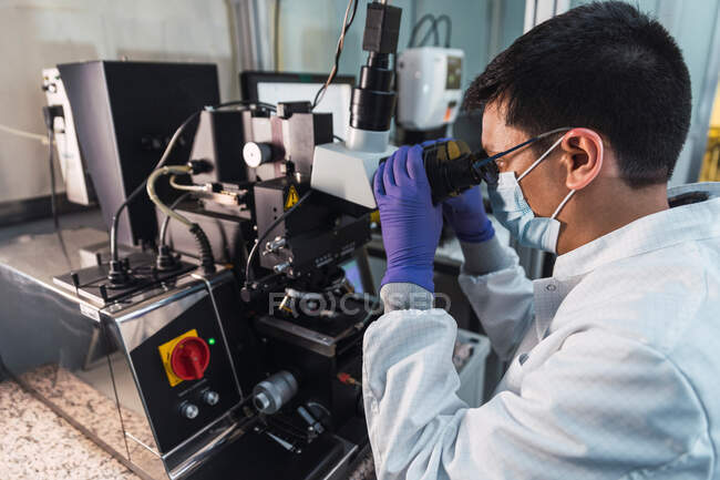 Visão lateral focada especialista masculino em luvas de jaleco e máscara facial examinando amostras através de poderosas lentes de microscópio enquanto trabalhava em laboratório moderno equipado — Fotografia de Stock
