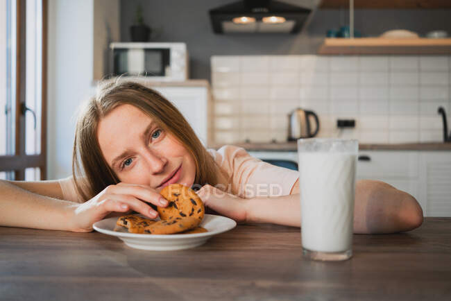 Junge Frau blickt in die Kamera, während sie am Tisch mit leckeren Haferflockenkeksen und einem Glas Milch zum Frühstück sitzt — Stockfoto