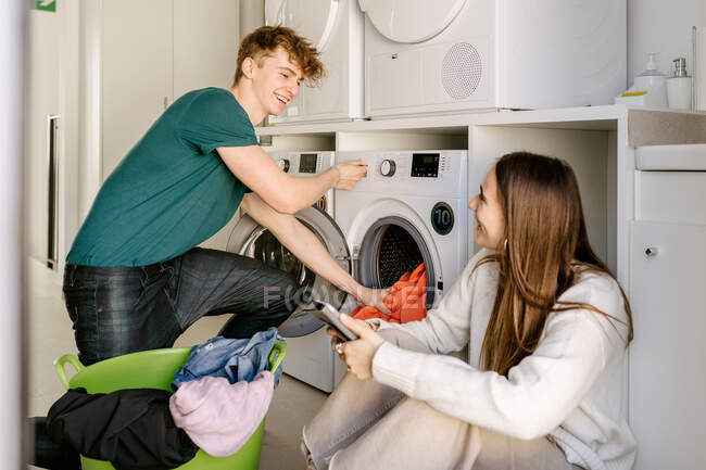 Весёлый молодой парень в повседневной одежде загружает одежду в стиральную машину и улыбается, общаясь с позитивной девушкой, используя смартфон в современной прачечной — стоковое фото