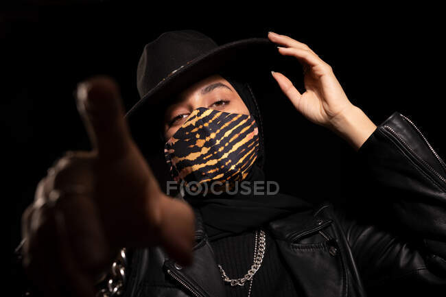Fiduciosa donna musulmana elegante in giacca di pelle alla moda maschera facciale e cappello toccare orlo e puntare la fotocamera mentre si guarda la fotocamera in studio scuro — Foto stock