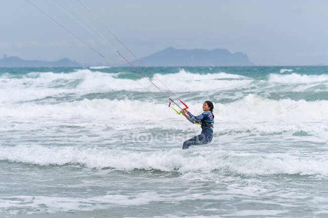 Aktive Sportlerin auf dem Kiteboard hält Kontrollstange beim Kitesurfen und schaut auf schäumenden Ozean weg — Stockfoto