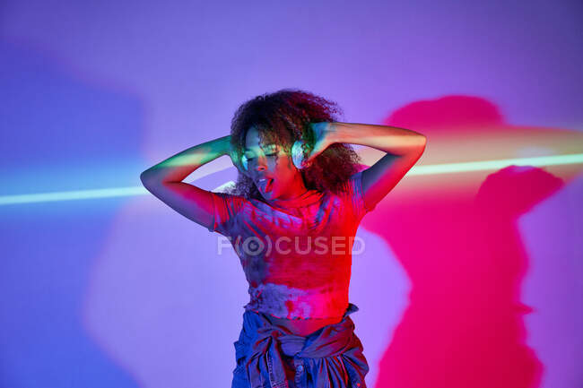 Full body African American female in shorts listening to favorite music in headphones and dancing looking away with tongue out in neon lights in studio — Stock Photo