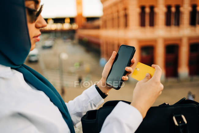 Vista lateral da cultura muçulmana elegante do sexo feminino pagando com cartão de plástico para a ordem durante as compras on-line via smartphone na cidade — Fotografia de Stock