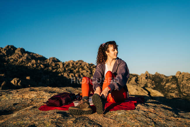 Full body of positive young female traveler with long curly hair in casual clothes and sunglasses smiling while tying laces of boots on rocky mountain slope on sunny day — Stock Photo