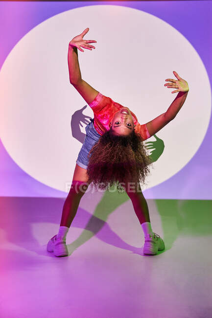 Гибкая афро-американская танцовщица в шортах загибается назад и смотрит в камеру, танцуя в студии в неоновых огнях — стоковое фото