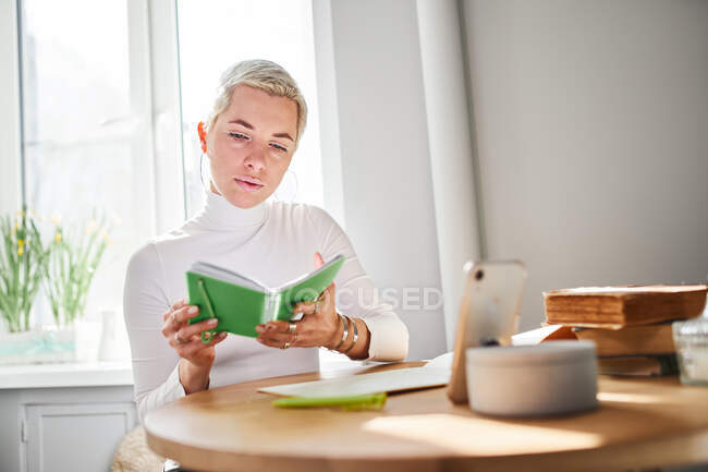 Astróloga adulta leyendo bloc de notas en el escritorio con smartphone y libros en casa a la luz del sol - foto de stock