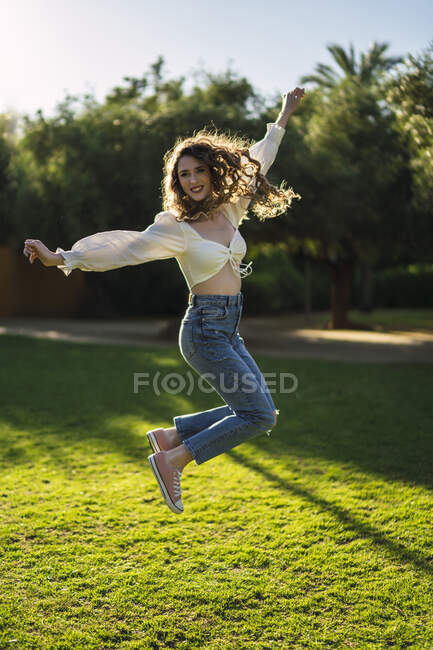 Стильна молода жінка з оголеним животом стрибає на пишному трав'янистому газоні в сонячному парку і дивиться на камеру — стокове фото