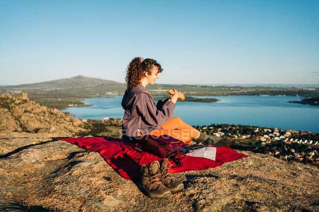 Vista lateral do jovem caminhante feliz em roupas casuais comendo sanduíche enquanto sentado em cobertor na colina rochosa sobre o mar contra o céu azul sem nuvens — Fotografia de Stock