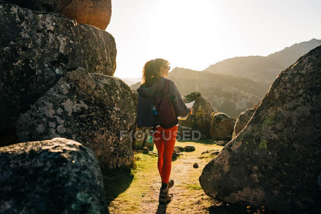 Обратный вид на неузнаваемую молодую женщину-исследователя с темными волосами в повседневной одежде, идущую среди скалистых валунов и читающую карту против безоблачного голубого неба — стоковое фото