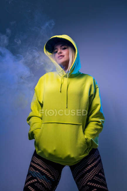 De baixo confiante hipster feminino com capuz fumar e cigarro no estúdio em fundo cinza e olhando para a câmera — Fotografia de Stock