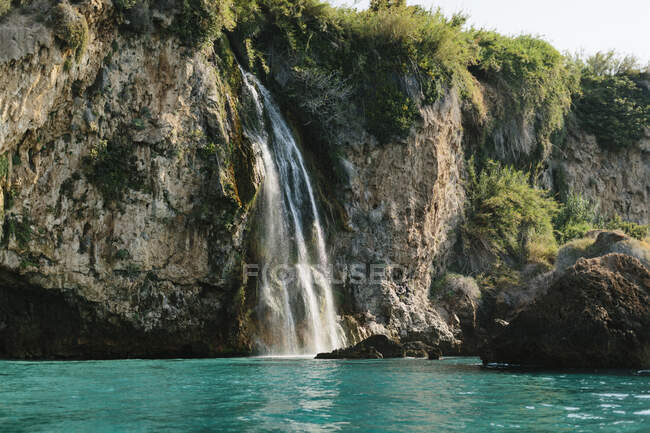 Increíble vista de la rápida cascada que cae de un acantilado áspero a una laguna ondulada de color turquesa en el soleado clima de verano en Málaga España - foto de stock