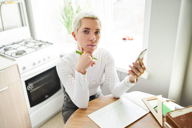 Женщина-астролог с линиями для рисования сотовых телефонов в бумажном альбоме за столом в светлом доме, глядя в камеру — стоковое фото