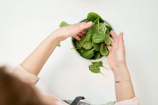 Colheita fêmea irreconhecível com folhagem de espinafre sobre tigela na mesa durante o processo de cozimento em casa — Fotografia de Stock
