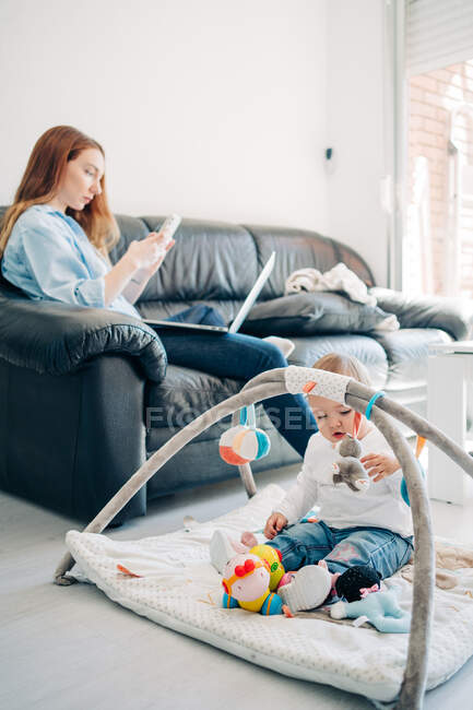 Орієнтована молода мати в повсякденному вбранні, переглядаючи смартфон і нетбук, сидячи на дивані біля чарівної маленької дитини, що грає з іграшками на підлозі у вітальні — стокове фото