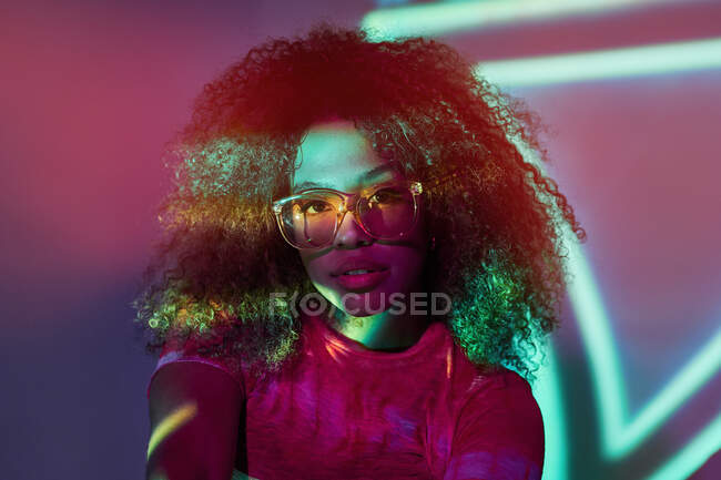 Портрет привабливої молодої афроамериканської жінки в стильних сонцезахисних окулярах, яка дивиться на камеру, стоячи в неонових вогні. — стокове фото