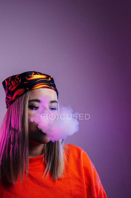 Vista lateral de la hembra fresca en traje de calle fumar cigarrillo electrónico y la exhalación de humo a través de la nariz en el fondo púrpura en el estudio con iluminación de neón rosa - foto de stock