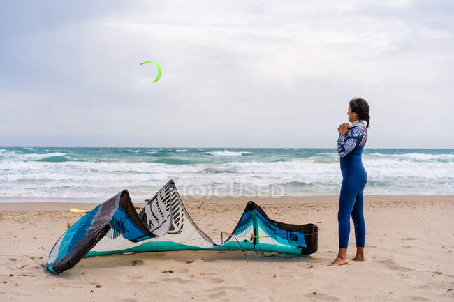 Vista lateral del kiter hembra descalzo en traje de neopreno de pie en la playa de arena del océano contra la cometa de energía bajo el cielo nublado - foto de stock