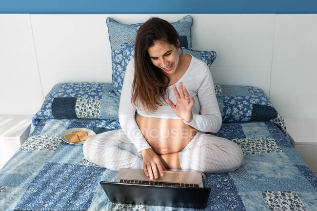 Сверху улыбающаяся беременная женщина говорит на видео-звонок на нетбуке и машет рукой, сидя на удобной кровати дома — стоковое фото