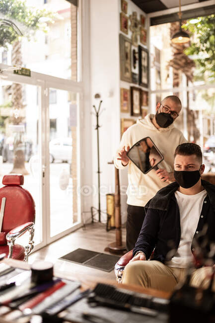 Peluquería masculina con espejo mostrando corte de pelo al cliente en máscara facial de tela mientras mira hacia adelante en la barbería durante la pandemia de COVID 19 - foto de stock