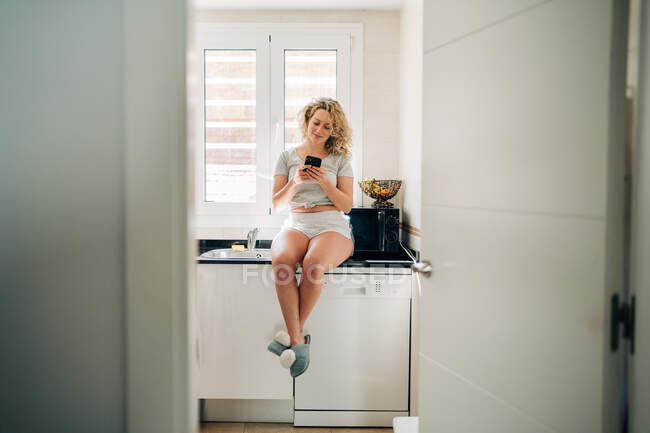Young content female in casual domestic wear browsing on smartphone while sitting on kitchen counter — Foto stock
