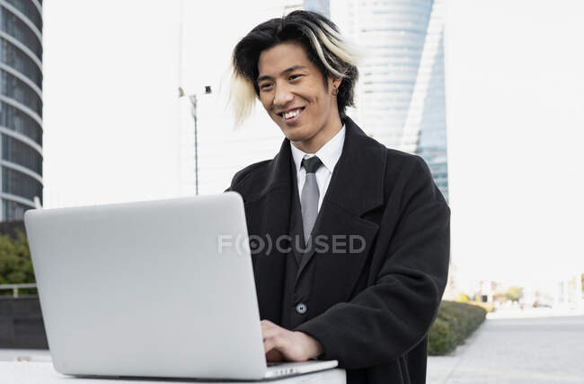 Giovane ben vestito felice etnico dirigente maschile con i capelli tinti che lavorano su netbook contro i moderni edifici della città — Foto stock