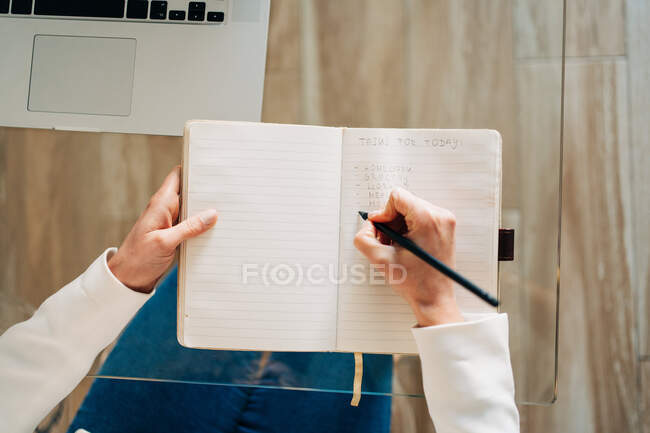 Зверху анонімна студентка з кучерявим волоссям у повсякденному одязі, що сидить за скляним столом і робить нотатки в копіювальній книзі, готуючись до іспитів, використовуючи ноутбук вдома — стокове фото