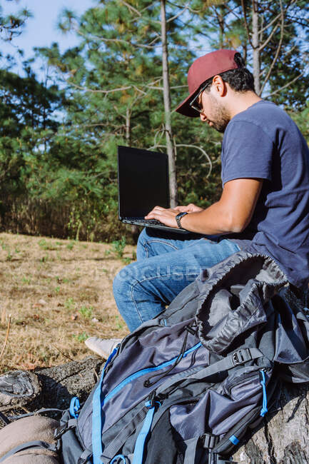 Desde abajo nómada sentado en piedra cerca de pertenencias y el uso de netbook mientras trabaja de forma remota - foto de stock