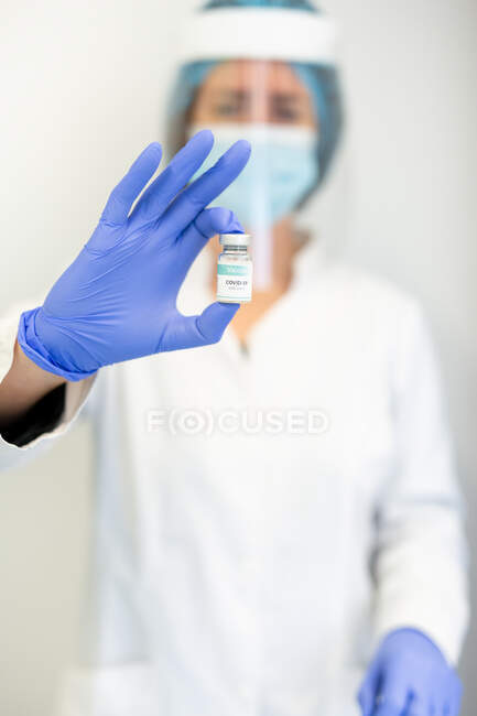 Doctora en guantes de látex y protector facial de pie con líquido químico en un vial de vidrio que se prepara para vacunar al paciente en la clínica durante el brote de coronavirus - foto de stock
