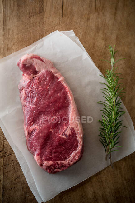 Overhead view of uncooked meat piece with rosemary leaves against baking paper on brown background — Stock Photo