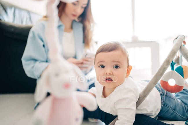 Adorable bebé en el suelo con juguetes mirando a la cámara mientras la madre irreconocible navega en el teléfono móvil en la sala de estar de luz - foto de stock