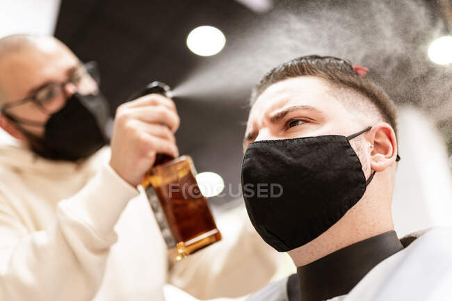 Estilista masculino en máscara textil rociando corte de pelo del cliente con líquido de la botella en la barbería - foto de stock