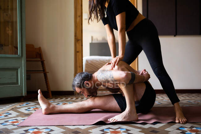 Side view of flexible male doing yoga in Pascimottanasana with help of crop personal instructor while sitting on mat at home — Stock Photo