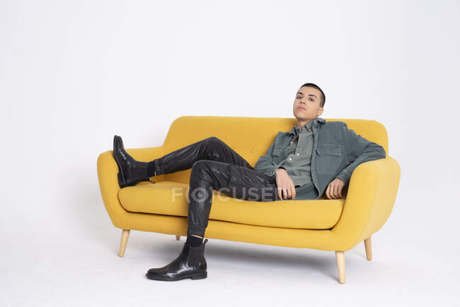 Молода красива чоловіча модель в модному одязі, що сидить на жовтому дивані на білому тлі в студії — стокове фото