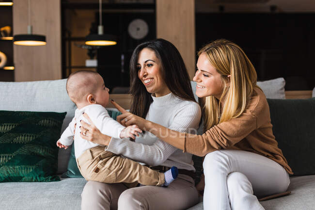 Cheerful couple of homosexual women playing with cute toddler while having fun and enjoying weekend together at home — Stock Photo