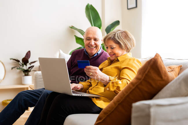 Joyeux couple d'âge mûr effectuant le paiement avec une carte en plastique pendant les achats en ligne via un ordinateur portable à la maison — Photo de stock