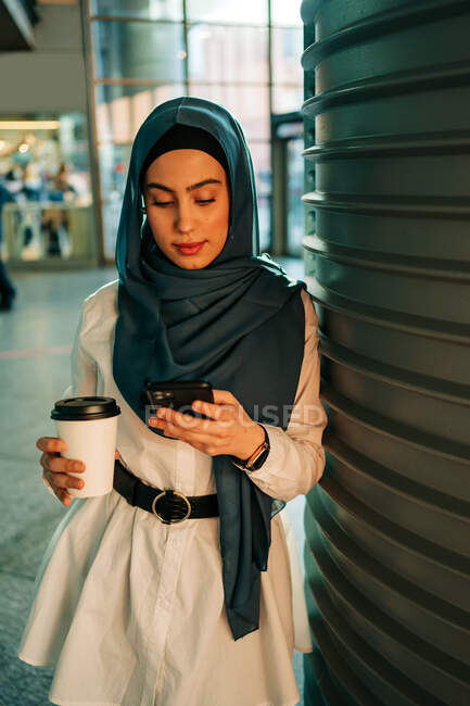 Ethnic female in hijab standing on station and browsing mobile phone while waiting for train — Stock Photo