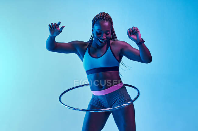 Весела афроамериканська спортсменка, яка займається активацією, обертальною обручкою під час тренування, дивлячись вниз у неоновому світлі. — стокове фото