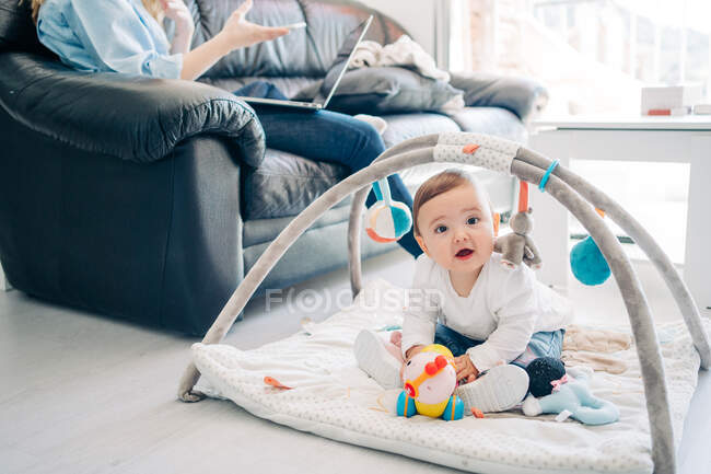 Recadré mère méconnaissable en tenue décontractée navigation sur smartphone et netbook assis sur canapé près adorable petit bébé regardant la caméra tout en jouant avec des jouets sur le sol dans le salon — Photo de stock