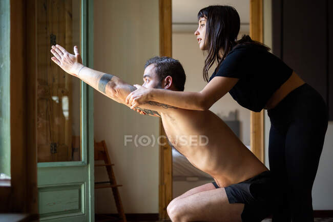 Vista lateral do macho concentrado em pé em Utkatasana e equilíbrio enquanto pratica ioga com a ajuda do instrutor pessoal feminino — Fotografia de Stock