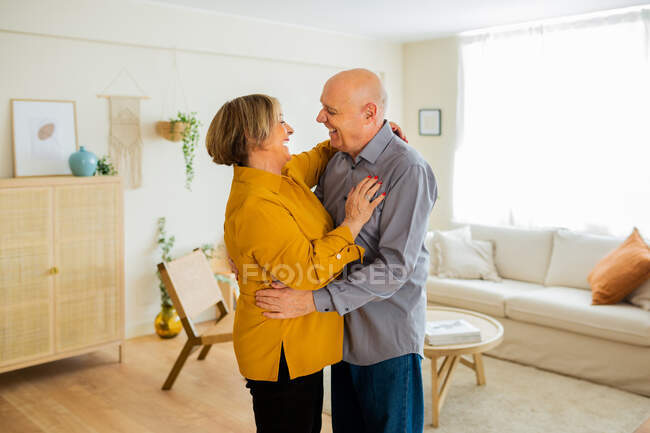 Glückliches Paar mittleren Alters, das sich zu Hause im Wohnzimmer umarmt und tanzt, während es sich zärtlich ansieht — Stockfoto