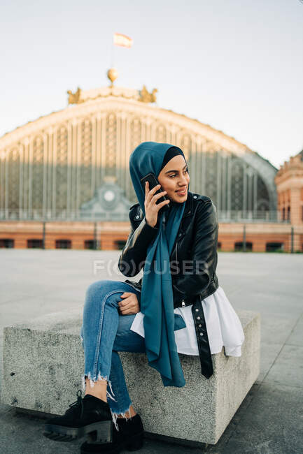 Mujer musulmana en pañuelo en la cabeza sentada en el banco mientras habla de teléfono móvil en la ciudad - foto de stock