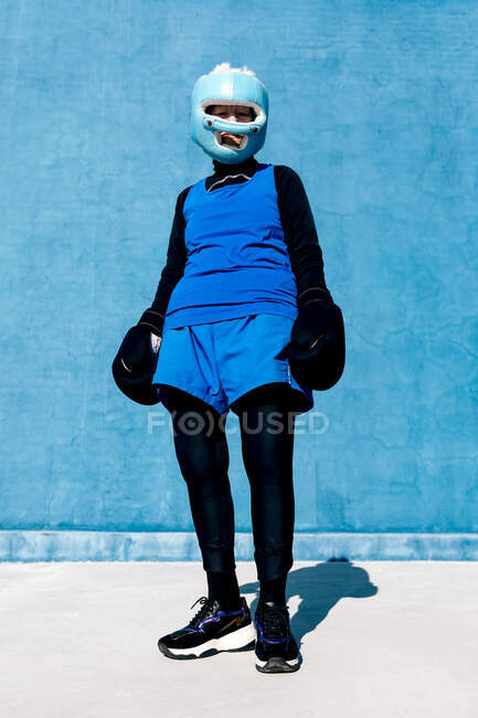 From below full length mature female in sportswear and boxing gloves standing with helmet against blue wall and looking at camera — Stock Photo