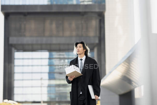 Jovem executivo étnico masculino de casaco com maquete de casa e netbook olhando para longe enquanto passeia na cidade — Fotografia de Stock
